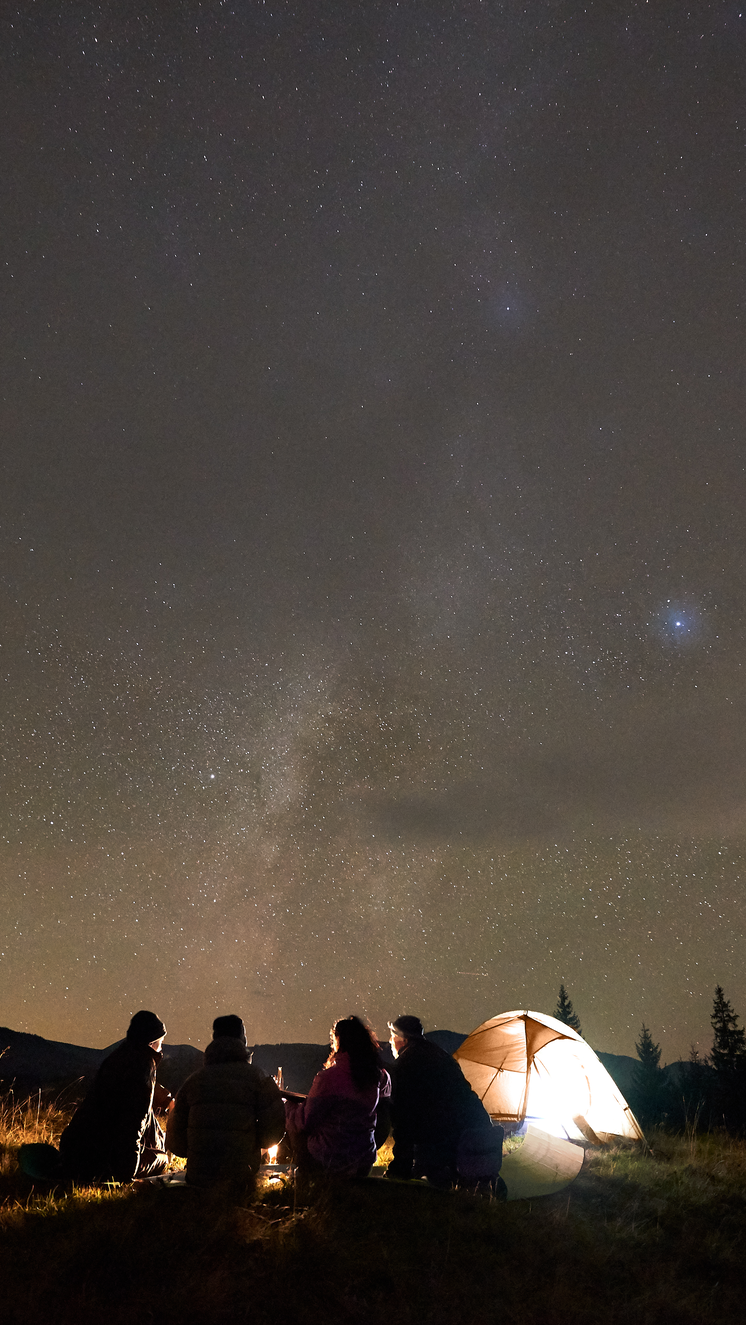 Campers sit in front of a small fire next to their tent, underneath a huge sky full of stars.